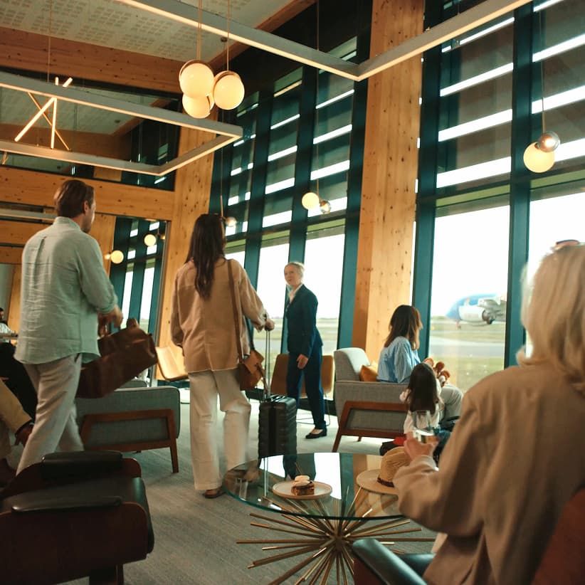 Travelers in an airport lounge with large windows, modern seating, and a plane visible outside. Some carry luggage, others sit and relax.
