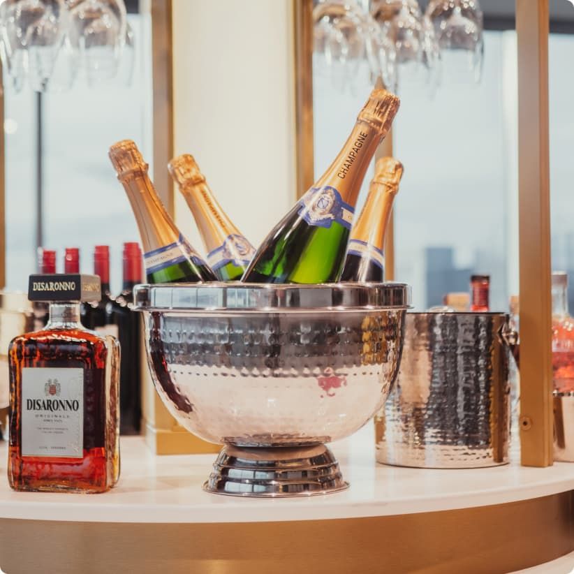 A metal ice bucket holds champagne bottles on a bar counter, surrounded by liquor bottles and glasses, with a cityscape visible in the background.