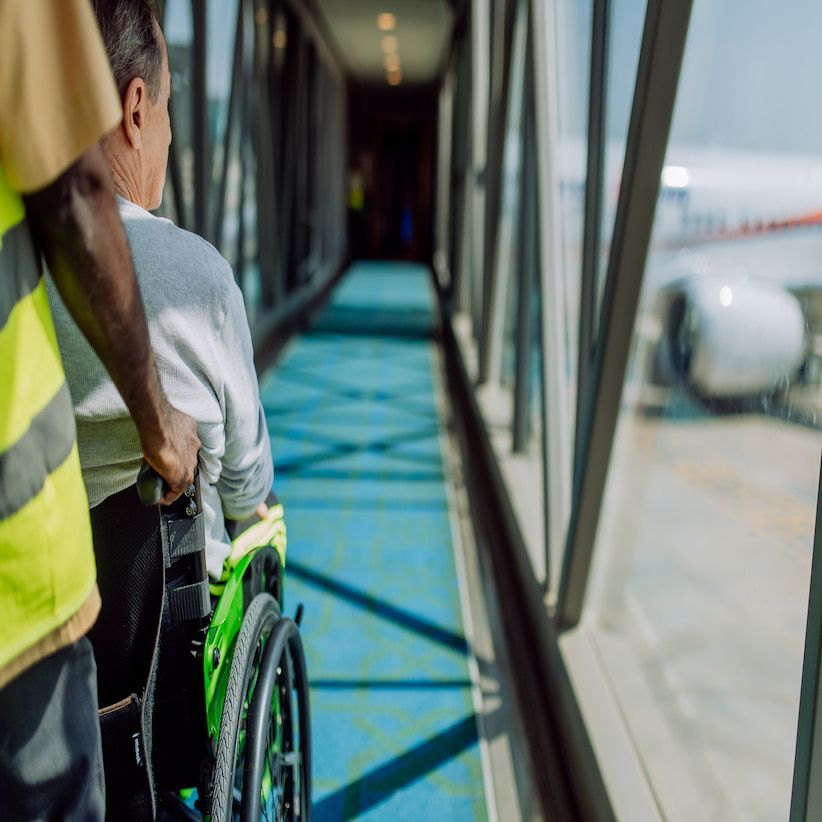 A person in a wheelchair is being assisted by an airport staff member through a glass-walled corridor, with an airplane visible outside.