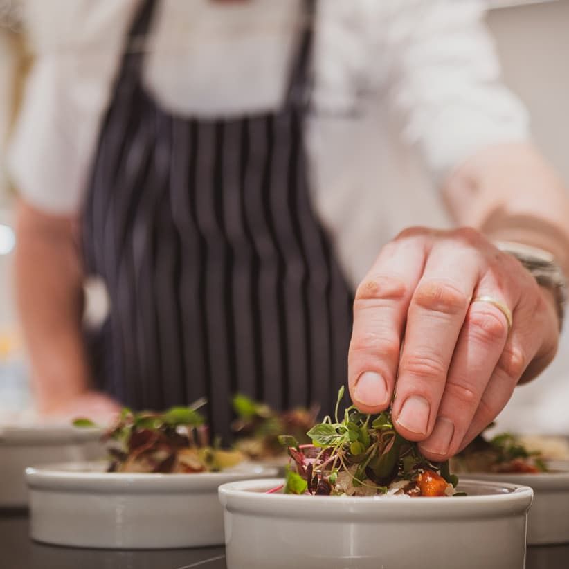 Chef garnishing dishes with fresh greens, wearing a striped apron. Close-up of hand and bowls with a blurred background.