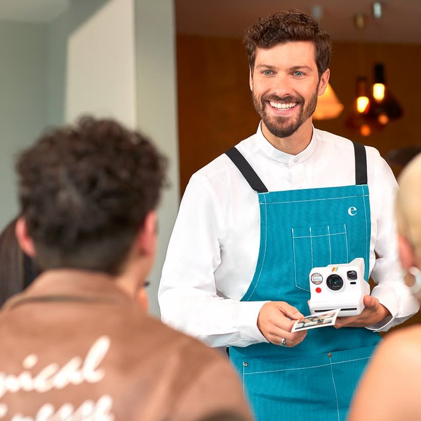 Smiling waiter in a teal apron shows a photo from an instant camera to customers in a restaurant setting.