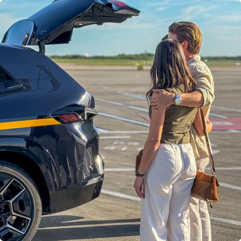 A couple embraces near a car with an open trunk on an airfield, facing the runway under a clear sky.