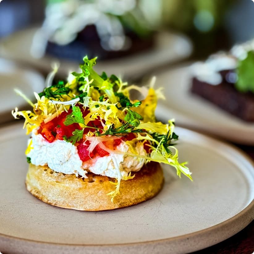 A gourmet dish featuring a crumpet topped with creamy cheese, frisée lettuce, diced red peppers, and herbs on a beige plate.