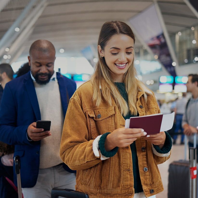 Woman smiling at passport and boarding pass, standing in airport line; man behind her checks his phone. Busy terminal background.