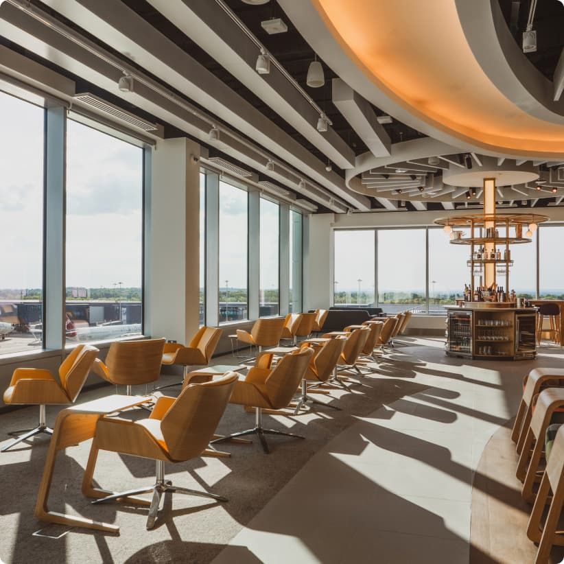 Modern airport lounge with wooden chairs, large windows, and a central circular bar. Natural light floods the space, creating a warm atmosphere.