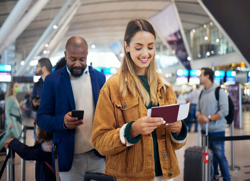 Woman smiling and holding a passport and boarding pass in an airport queue, with a man checking his phone behind her. Blurred people in background.