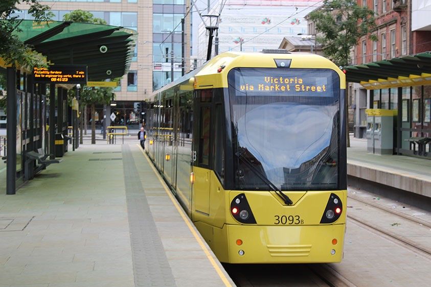Yellow tram at a city station with "Victoria via Market Street" displayed, surrounded by modern buildings and green shelters.