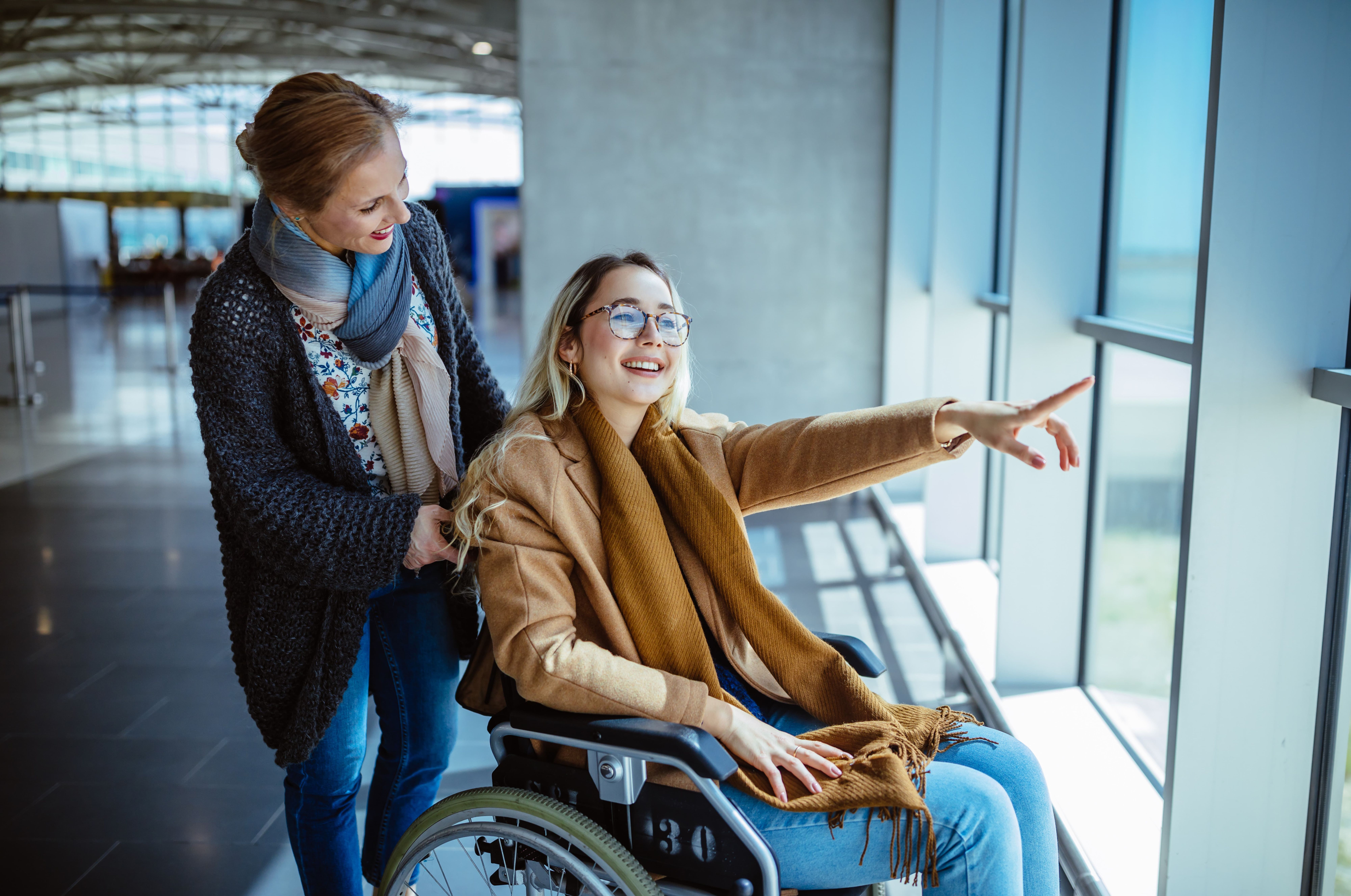 A woman in a wheelchair, smiling and pointing out a window, is accompanied by another woman in an airport.
